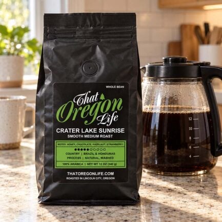 Crater Lake Sunrise Smooth Medium Roast whole bean coffee in a black 12 oz That Oregon Life bag beside a glass coffee pot on a sunlit kitchen counter.