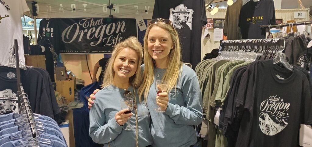 Two smiling visitors holding glasses at the That Oregon Life clothing booth
