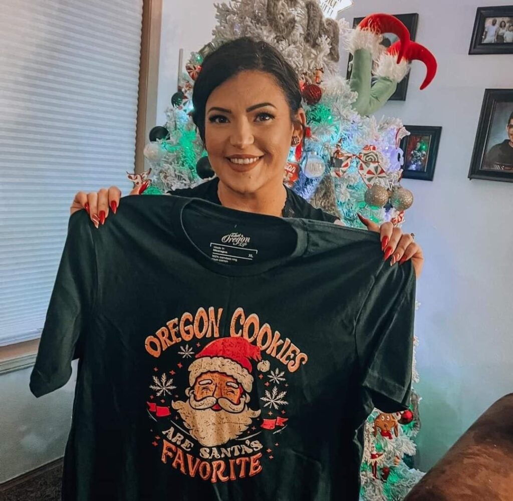 Woman holding an Oregon Cookies Are Santa’s Favorite shirt beside a Christmas tree