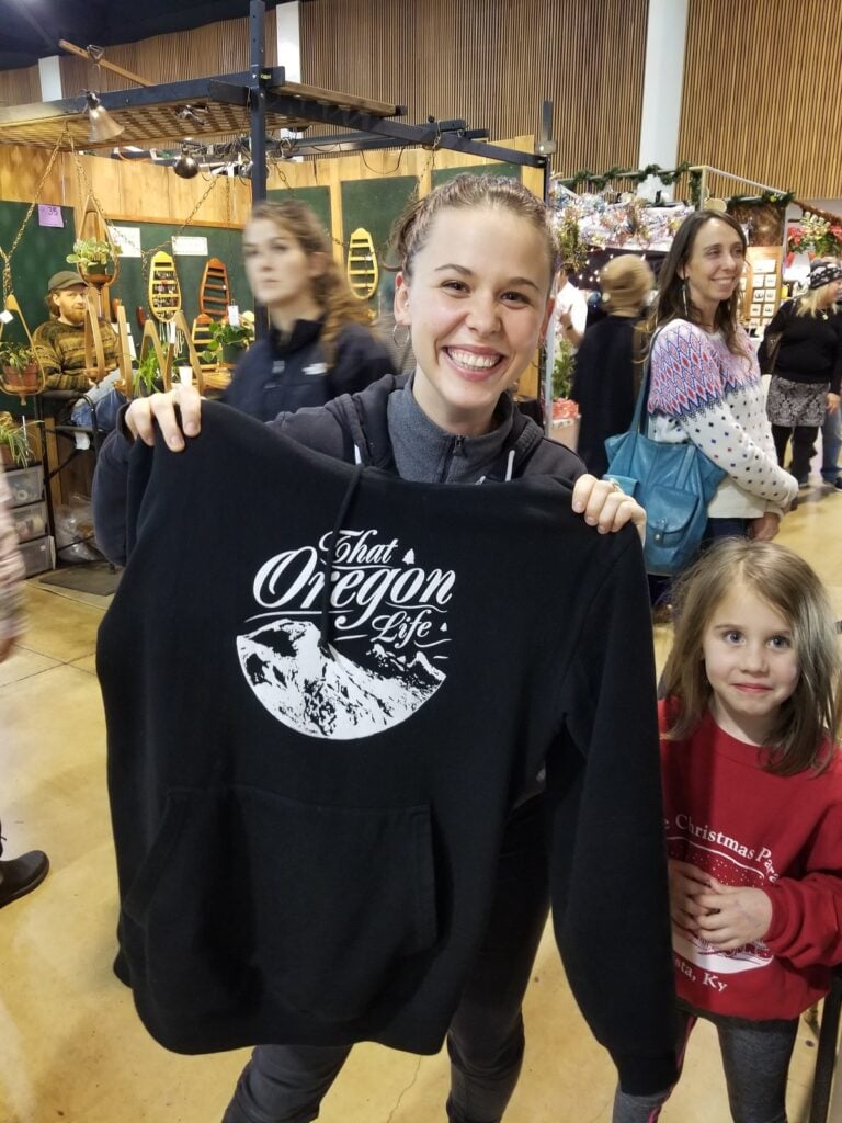 Smiling woman holding a black That Oregon Life mountain hoodie at a local market