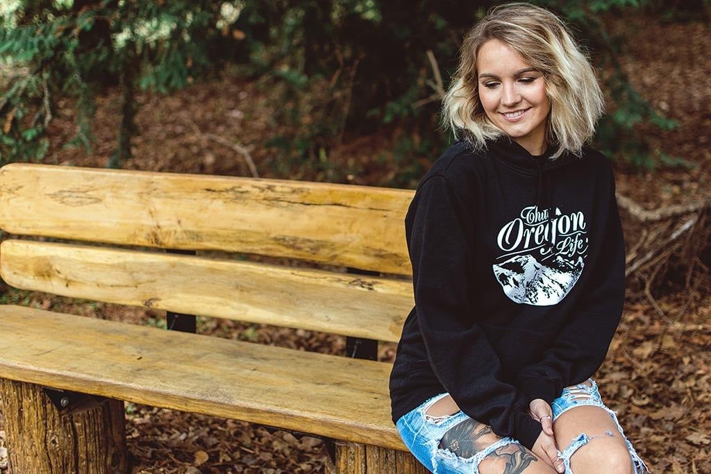 Woman wearing a black That Oregon Life mountain hoodie on a wooden bench in the forest